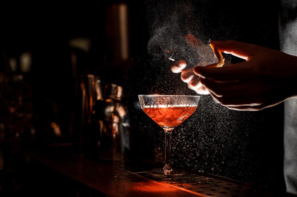 Bartender spritzt mit einem Orangenschnitz in ein Cocktail Glas